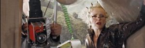 Still from Harry Potter ad the Goblet of Fire: Middle-aged blond woman with cat-eye glasses (Rita Skeeter) with quill and pad of paper.